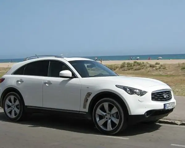 Białe infiniti auto zaparkowane przy plaży, z widokiem na morze i ludzi odpoczywających na piasku.