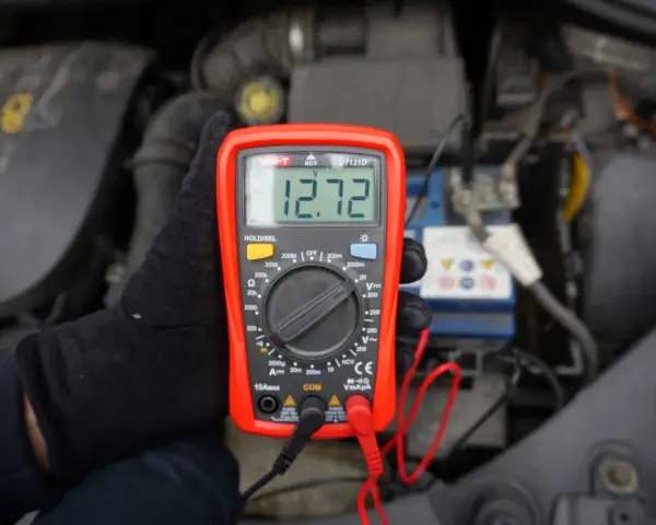 Ręka w czarnej rękawiczce trzyma multimetr pokazujący 12.72V. To idealne minimalne napięcie akumulatora do uruchomienia.