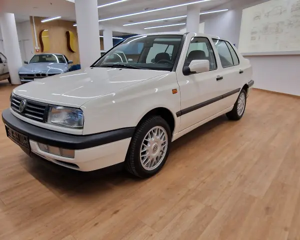 Biały Volkswagen Vento z lat 90. stoi na drewnianej podłodze w salonie samochodowym.