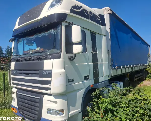 Ciężarówka DAF XF 105 z niebieską plandeką stoi na trawie, częściowo zasłonięta przez krzaki.