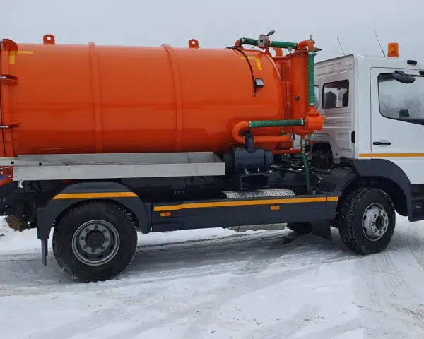 Pomarańczowy pojazd asenizacyjny z beczką i zielonymi wężami stoi na zaśnieżonej drodze.