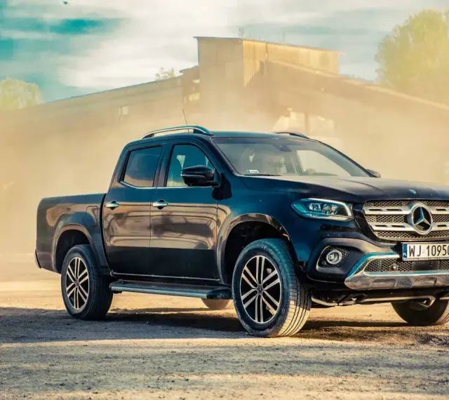 Czarny Mercedes pickup z przyciemnianymi szybami, zaparkowany na piaszczystym podłożu, w tle przemysłowy budynek i unoszący się pył.