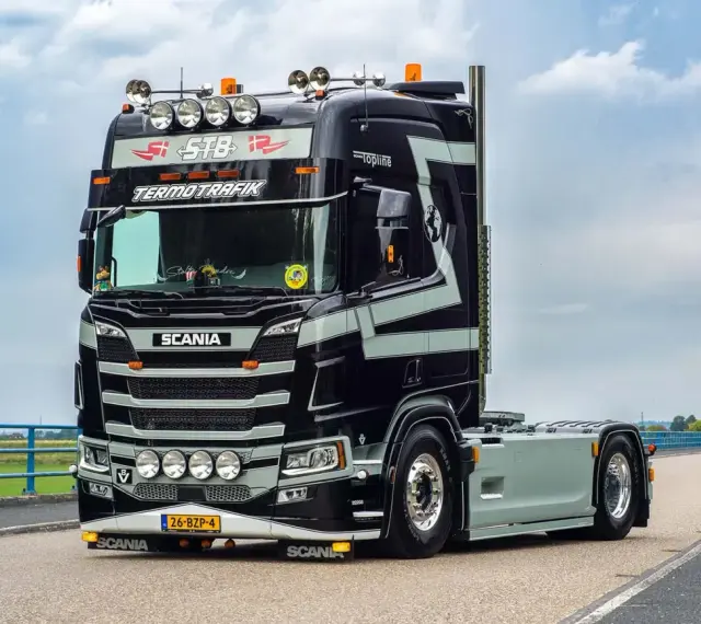 Czarna Scania V8 z szarymi pasami, z napisem "TERMOTRAFIK" i licznymi światłami, gotowa do trasy.