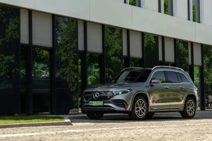 Nowy Mercedes 4x4, elegancki SUV, zaparkowany przed nowoczesnym budynkiem z zielonymi odbiciami drzew w szybach.