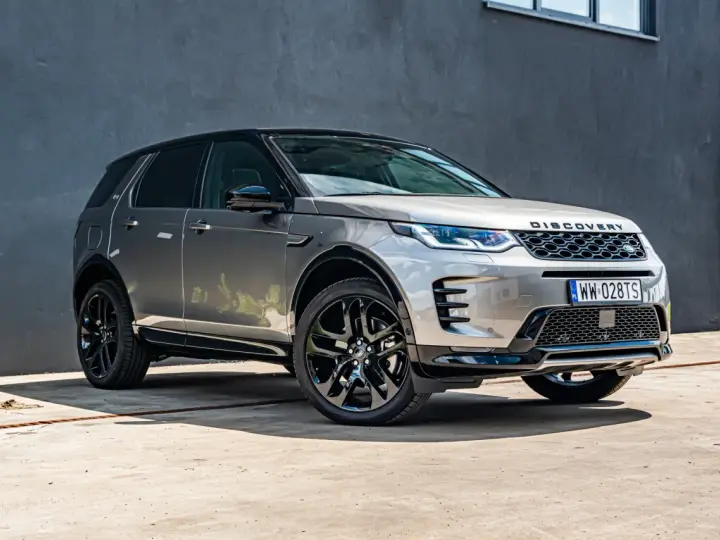 Srebrny Land Rover Discovery Sport z czarnym dachem, zaparkowany przed szarą ścianą.