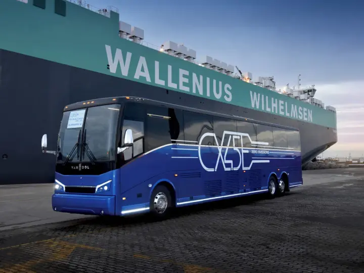 Niebieski autokar Van Hool CX45 z napisem "ZERO EMISSIONS" stoi przy ogromnym statku Wallenius Wilhelmsen.