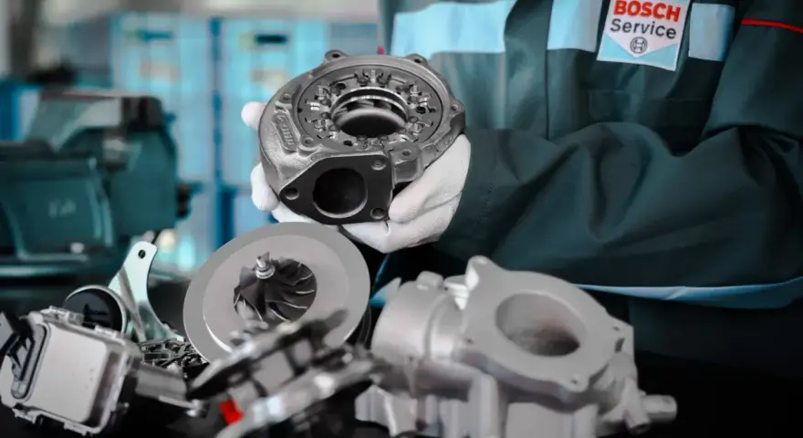 Mechanik w białych rękawiczkach prezentuje części turbosprężarki, sugerując domowe ustawienie sztangi turbiny.