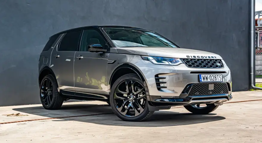 Srebrny Land Rover Discovery Sport z czarnym dachem, zaparkowany przed szarą ścianą.