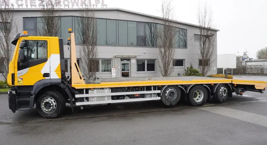 Żółty Iveco Stralis z platformą transportową stoi przed budynkiem.