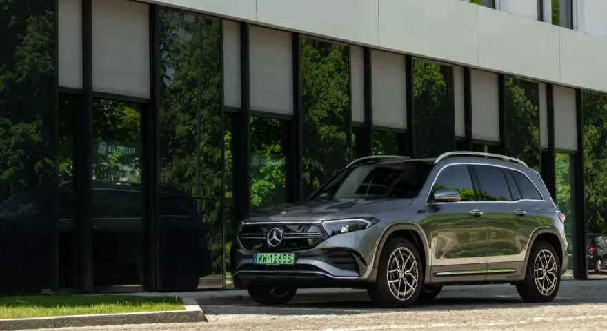 Nowy Mercedes 4x4, elegancki SUV, zaparkowany przed nowoczesnym budynkiem z zielonymi odbiciami drzew w szybach.