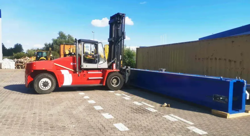 Czerwony wózek widłowy przygotowuje się do przeładunku długiego, niebieskiego elementu. To przykład sprawnego cross dockingu.