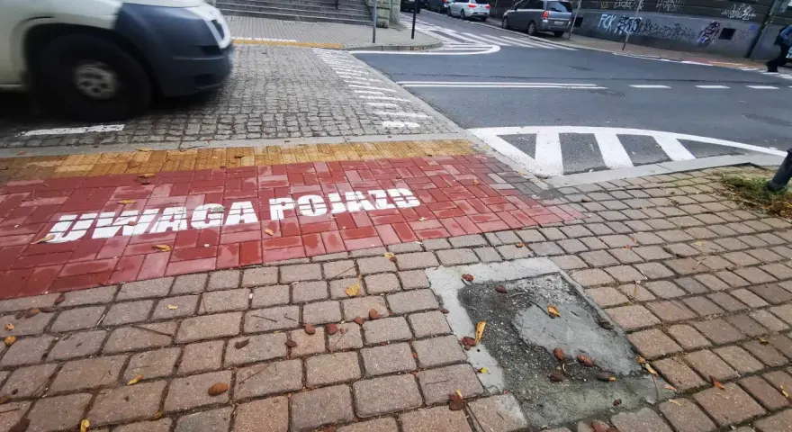 Czerwony pas na chodniku z napisem "UWAGA POJAZDY" informuje o przejściu sugerowanym. W tle widać samochody i schody.