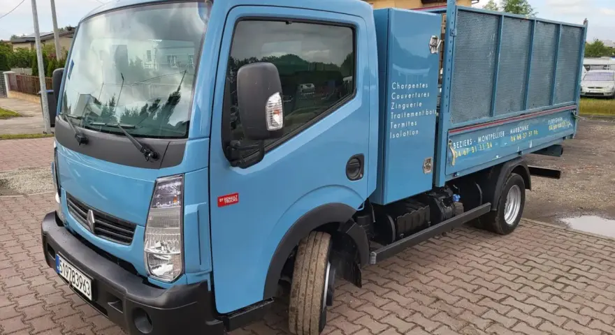 Niebieska ciężarówka Renault Maxity z otwartą skrzynią ładunkową, zaparkowana na brukowanym podjeździe.