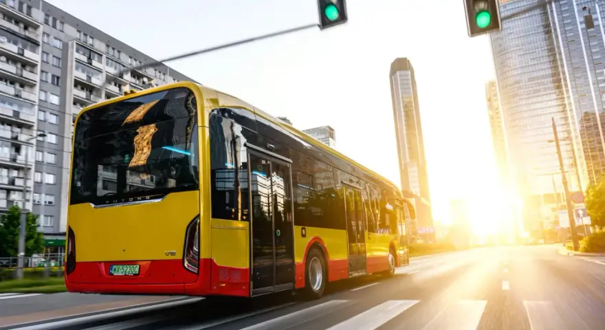 Żółto-czerwony autobus elektryczny Yutong jedzie przez miasto w blasku zachodzącego słońca.