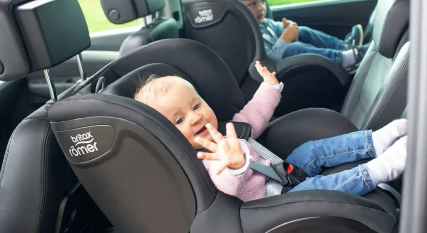 Uśmiechnięte niemowlę w foteliku Britax Römer, obok starsze dziecko. Od kiedy dziecko może jeździć z przodu? To pytanie nurtuje wielu rodziców.
