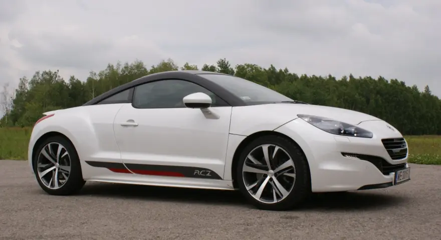 Biały Peugeot RCZ z czarnym dachem i sportowymi akcentami na progu, zaparkowany na tle zielonego lasu.
