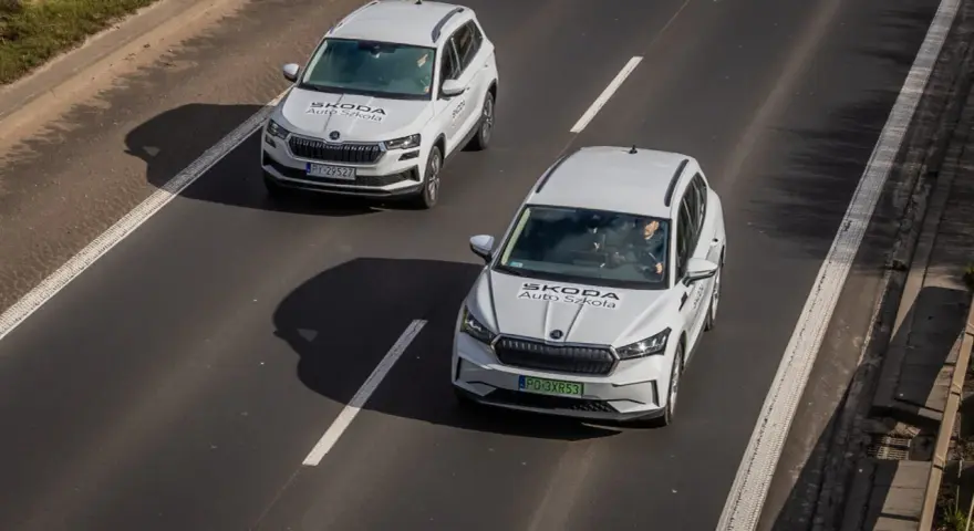 Dwa białe samochody Skoda Auto Szkoła jadą po drodze, gdzie obowiązuje ruch lewostronny.