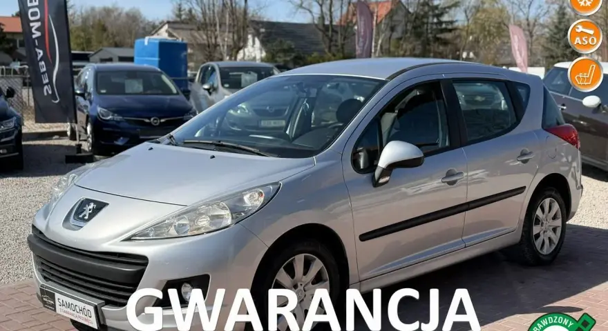 Srebrny Peugeot 207 1.4 benzyna na placu samochodowym, z napisem "GWARANCJA".