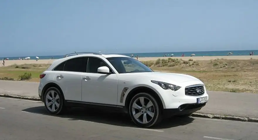 Białe infiniti auto zaparkowane przy plaży, z widokiem na morze i ludzi odpoczywających na piasku.