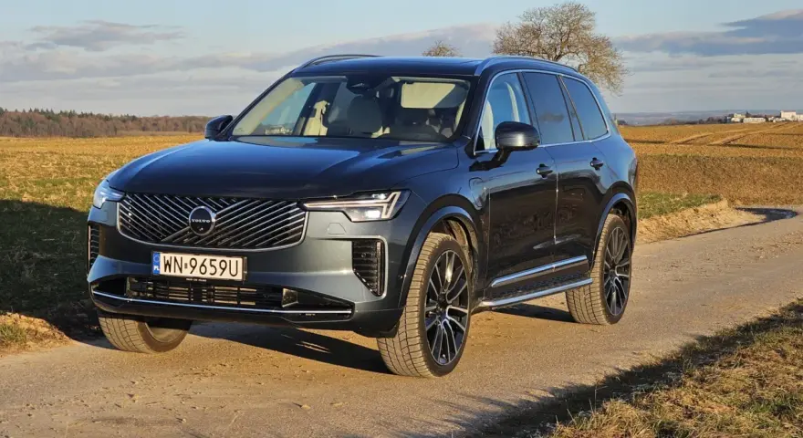 Ciemnoszary Volvo XC90 na polnej drodze, z widokiem na pola i drzewa w tle.