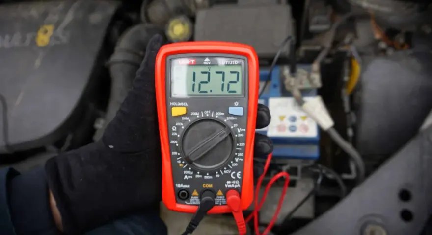 Ręka w czarnej rękawiczce trzyma multimetr pokazujący 12.72V. To idealne minimalne napięcie akumulatora do uruchomienia.