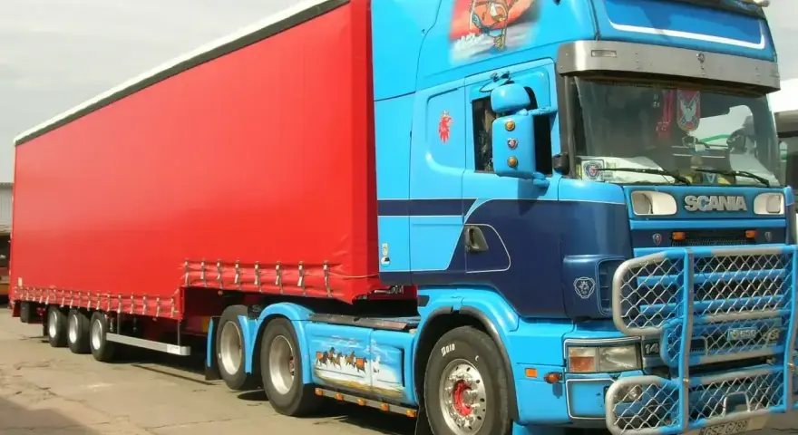 Naczepa jumbo w jaskrawym kolorze czerwonym, ciągnięta przez niebieską Scanię z malowidłami.
