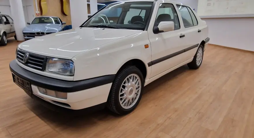 Biały Volkswagen Vento z lat 90. stoi na drewnianej podłodze w salonie samochodowym.