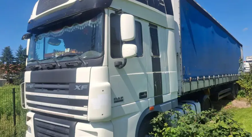 Ciężarówka DAF XF 105 z niebieską plandeką stoi na trawie, częściowo zasłonięta przez krzaki.
