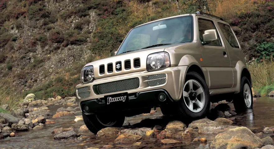 Złoty Suzuki Jimny pokonuje kamienisty strumień. Sprawdź suzuki jimny cena i możliwości terenowe tego kultowego auta.