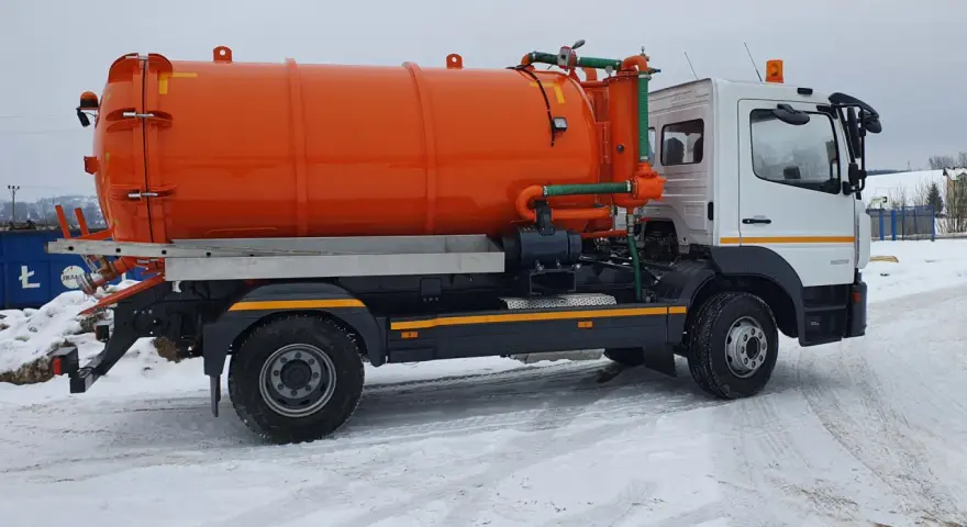 Pomarańczowy pojazd asenizacyjny z beczką i zielonymi wężami stoi na zaśnieżonej drodze.