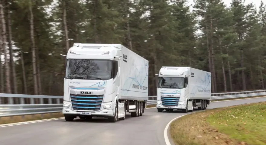 Dwa białe elektryczne DAF XG Plus z naczepami jadące po krętej drodze wśród drzew.
