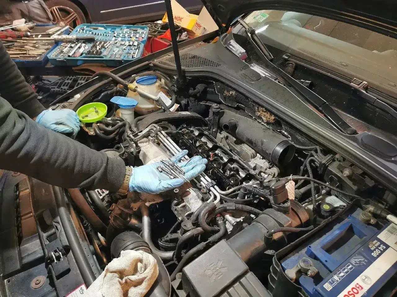 Mechanik w niebieskich rękawiczkach trzyma świece żarowe, przygotowując się do sprawdzenia ich stanu. W tle widać skrzynkę z narzędziami.