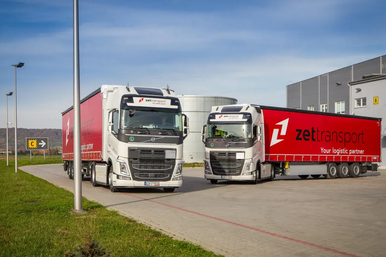 Dwa białe Volvo z czerwonymi naczepami Zetttransport, symbolizujące spedycję i logistykę.