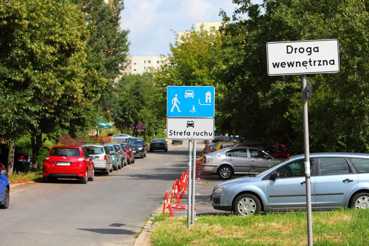 Znaki drogowe informują o wjeździe do strefy ruchu. Samochody stoją zaparkowane wzdłuż ulicy.