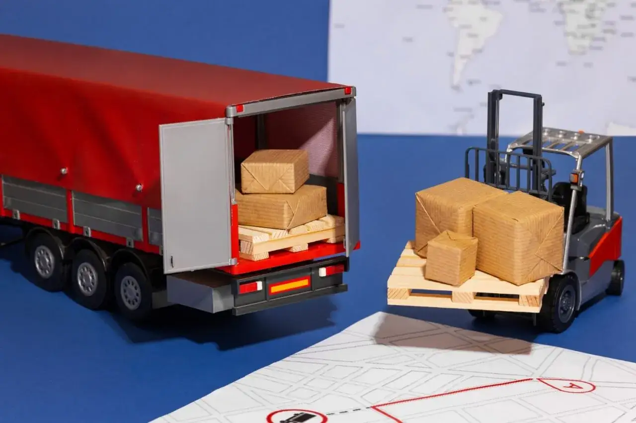 Miniaturowy wózek widłowy z paczkami zbliża się do otwartej naczepy ciężarówki. To idealny obrazek dla kogoś, kto planuje transport, może nawet z wykorzystaniem karnetu ATA.