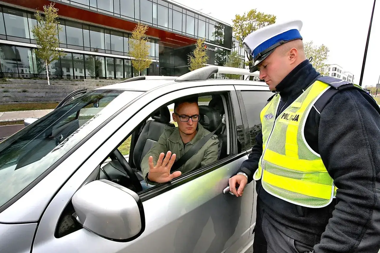 Policjant wręcza kierowcy mandat za brak przeglądu. Kierowca podnosi rękę w geście protestu.