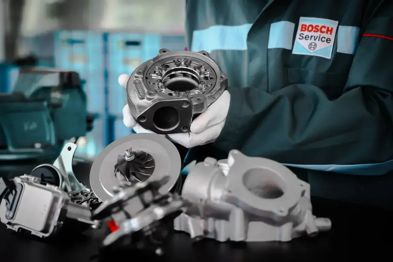 Mechanik w białych rękawiczkach prezentuje części turbosprężarki, sugerując domowe ustawienie sztangi turbiny.