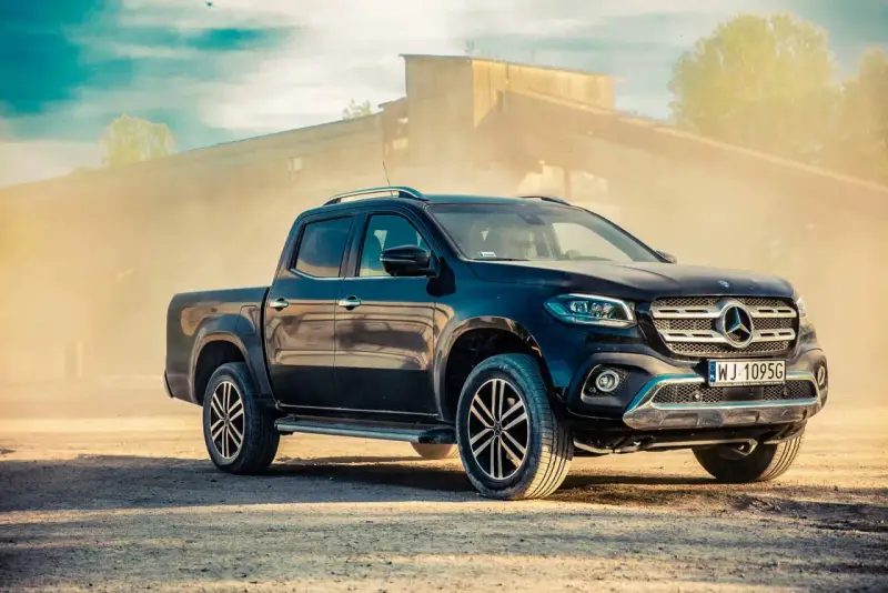Czarny Mercedes pickup z przyciemnianymi szybami, zaparkowany na piaszczystym podłożu, w tle przemysłowy budynek i unoszący się pył.
