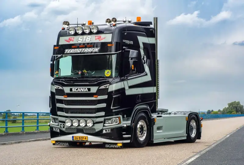 Czarna Scania V8 z szarymi pasami, z napisem "TERMOTRAFIK" i licznymi światłami, gotowa do trasy.